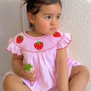 May include: A pink and white gingham baby dress with embroidered strawberries. The dress has short sleeves and a smocked bodice. The baby is wearing red hair bows.