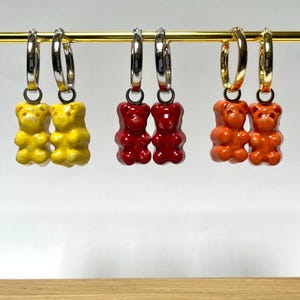 May include: Three pairs of gummy bear earrings in yellow, red, and orange. Each pair of earrings features a small, colorful gummy bear charm hanging from a silver or gold hoop earring. The earrings are displayed on a gold bar.