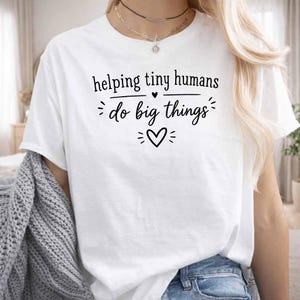 Puede incluir: Camiseta blanca con el texto negro "helping tiny humans do big things". El diseño incluye un símbolo de corazón. La camiseta es de estilo casual.