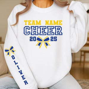 Könnte beinhalten: Weißes Sweatshirt mit der Aufschrift "TEAM NAME CHEER 2025" in blauen und gelben Buchstaben. Eine Schleifengrafik befindet sich mittig unter dem Text. Der Ärmel zeigt eine Schleifengrafik und den Namen "OLIVER" in Blau.
