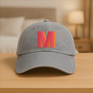 Op de afbeelding: Een grijze baseballpet met een gebogen klep. De voorkant toont een groot, geborduurd "M" in roze, oranje en geel. De pet is gemaakt van een zachte stof en heeft een verstelbare band.