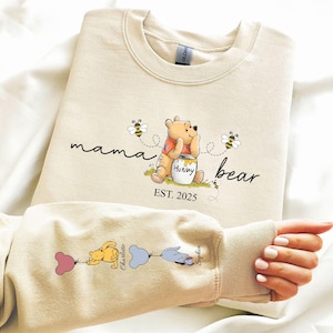 Könnte beinhalten: Ein beiger Pullover mit dem Text "mama bear est. 2025" und einem Cartoon von Winnie Puuh, der einen Honigtopf hält. Der Ärmel hat einen Cartoon von Winnie Puuh mit zwei Ballons, auf denen "Charlotte" und "Sophia" steht.