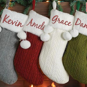 Puede incluir: Cuatro calcetines de Navidad personalizados con pompones. Los calcetines son grises, rojos, blancos y verdes con ribete de piel blanca y los nombres "Kevin", "Ameli", "Grace" y "Daniel" bordados en rojo.
