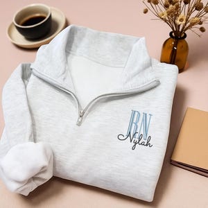 Może przedstawiać: Jasnoszara bluza z zamkiem błyskawicznym do połowy szyi z wyhaftowanymi literami "RN" nad imieniem "Nylah" w ozdobnej czcionce. Bluza jest złożona na różowej powierzchni, z filiżanką kawy i małym wazonem w tle.