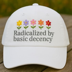Op de afbeelding: Witte baseballcap met geborduurde bloemen en de tekst "Radicalized by basic decency". De bloemen zijn geel, paars, roze, oranje en rood. De cap heeft een gebogen klep en een knoop bovenop.