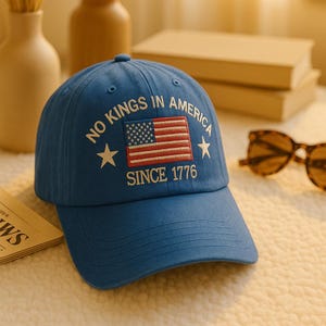 Può includere: Un cappellino da baseball blu con la scritta "NO KINGS IN AMERICA SINCE 1776" e un disegno della bandiera americana. Il cappellino ha una visiera curva e una classica costruzione a sei pannelli. Il testo e la bandiera sono ricamati in bianco e rosso.