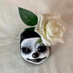 Pu&ograve; includere: Un vaso in ceramica bianca a forma di viso da clown con occhi neri, naso nero e bocca nera. Una rosa bianca con foglie verdi &egrave; posizionata sopra il vaso.