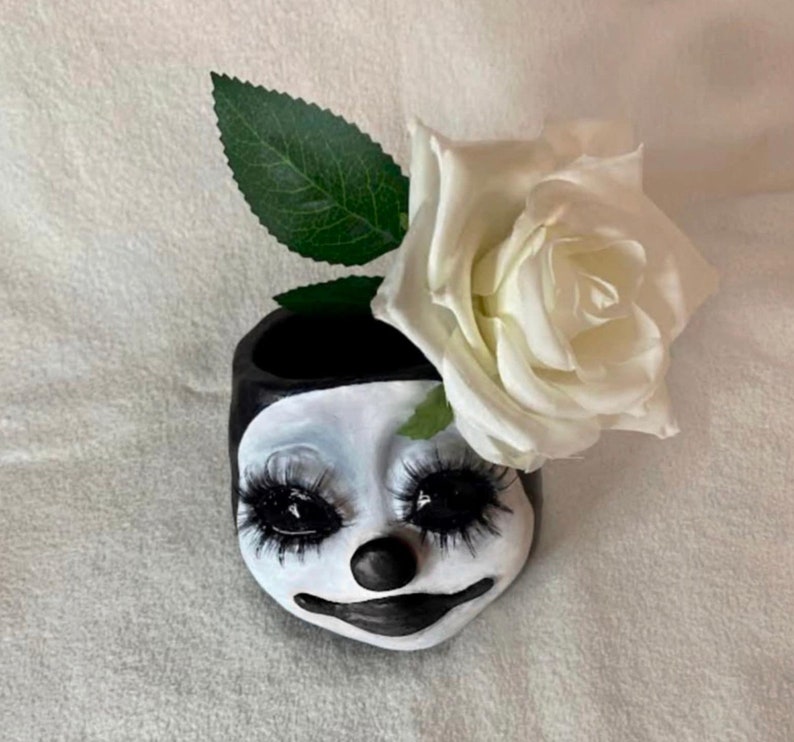 Pu&ograve; includere: Un vaso in ceramica bianca a forma di viso da clown con occhi neri, naso nero e bocca nera. Una rosa bianca con foglie verdi &egrave; posizionata sopra il vaso.