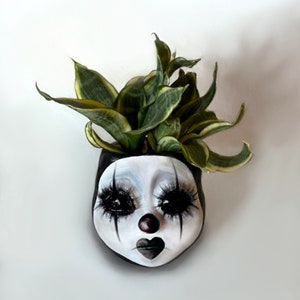 May include: A unique, hand-painted planter shaped like a clown's face, featuring a black and white color scheme with exaggerated eyelashes and a heart-shaped mouth. The planter is filled with green and yellow snake plants.