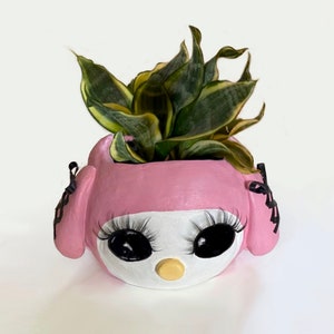 May include: A pink and white ceramic planter shaped like a cartoon character's face, with large black eyes, long eyelashes, and a yellow nose. The planter holds a green and yellow plant.