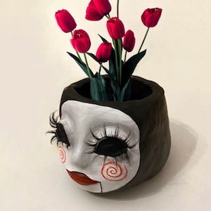 May include: A ceramic planter shaped like a doll's head with black hair, white skin, red lips, and black eyelashes. The doll's face has a spiral pattern painted on the cheeks. The planter is filled with red tulips.