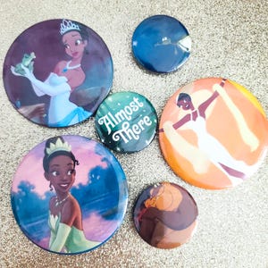May include: Five circular pinback buttons featuring animated characters. One shows a woman in a white gown holding a frog, another a woman in a green dress, one with the text Almost There, one with a dark blue character, and one with a brown creature.