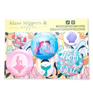 May include: A set of five circular pin-back buttons featuring The Little Mermaid characters and themes, including a crab, a couple in a boat, a silhouette, a mermaid tail, and a seashell, displayed on a card labeled Glass Slippers & coffee.