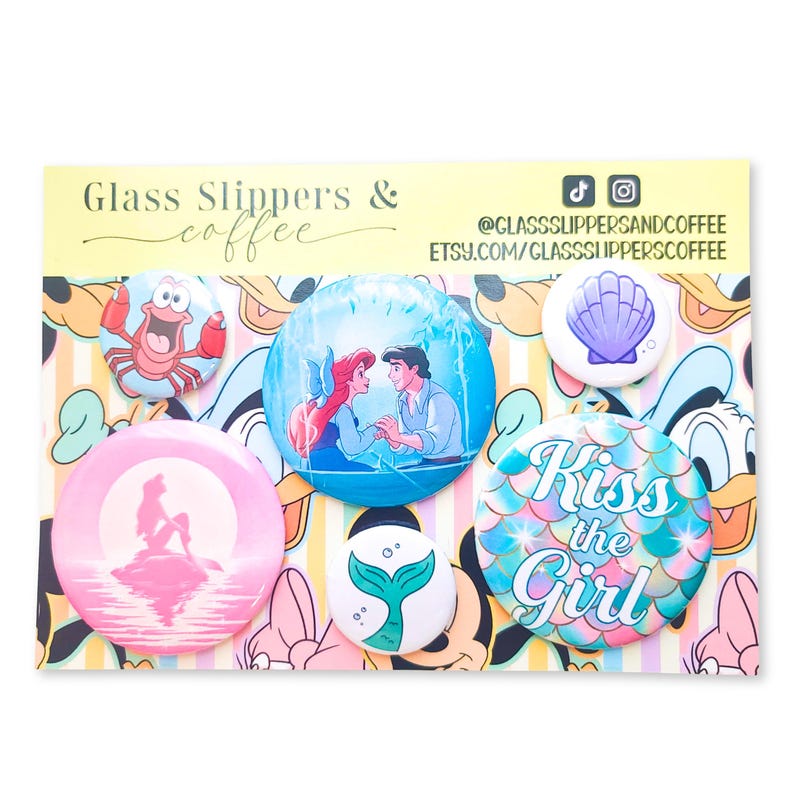 May include: A set of five circular pin-back buttons featuring The Little Mermaid characters and themes, including a crab, a couple in a boat, a silhouette, a mermaid tail, and a seashell, displayed on a card labeled Glass Slippers & coffee.