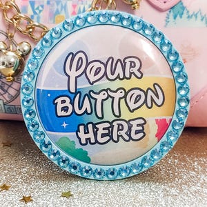 May include: A round, light blue button with the text "Your Button Here" in a whimsical font. The button is surrounded by a ring of blue rhinestones. The background of the button has a colorful, striped design.