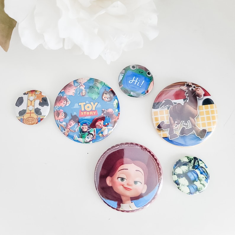 May include: A collection of six circular pin-back buttons featuring various designs. One button displays the Toy Story logo with characters, another shows a brown horse, one features a character with red hair, and others show patterns.