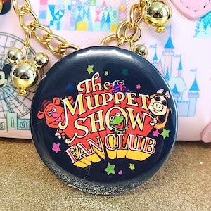 May include: A round, dark blue button with the words "The Muppet Show Fan Club" in orange and yellow. The button features cartoon images of Muppet characters and colorful stars. A gold chain and Mickey Mouse charm are visible.