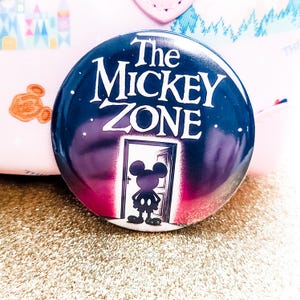 May include: A round button with the text "The MICKEY ZONE" in white, over a dark blue background with a silhouette of Mickey Mouse in a doorway. The button is on a gold glitter surface.