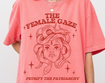 May include: A coral pink oversized t-shirt with a red graphic print of a woman's face with snakes for hair. The text "THE FEMALE GAZE" and "PETRIFY THE PATRIARCHY" are printed on the shirt.