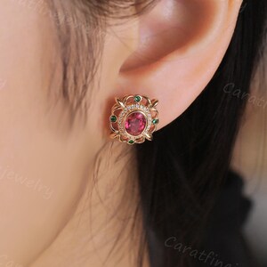 May include: Rose gold stud earrings featuring a central pink gemstone, encircled by smaller green and clear stones. The earrings have an ornate, floral design. The earrings are displayed on an ear.