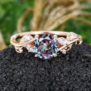 May include: Rose gold ring with a round alexandrite gemstone that changes colour. The ring has a floral design with small, clear accent stones. The ring is set on a dark, textured surface.