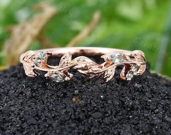 May include: A delicate rose gold band ring with a leafy design and small green gemstones. The ring is set on a dark gray background.