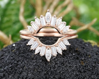 May include: A rose gold wedding band set with a delicate, curved, marquise-shaped diamond band. The band is adorned with a row of smaller diamonds that create a sunburst effect.