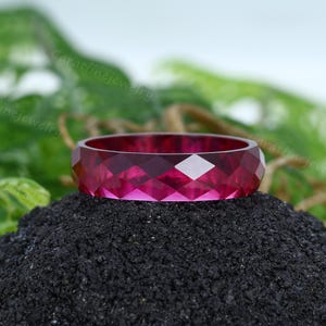 Puede incluir: Un anillo facetado de color magenta. El anillo está colocado sobre una superficie oscura y texturizada, con follaje verde en el fondo. La superficie del anillo está cortada con múltiples facetas, creando un patrón geométrico.