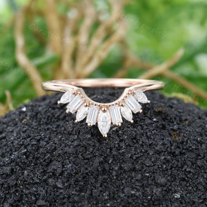 Könnte beinhalten: Ein roségoldener Ehering mit einem zarten, geschwungenen Design. Der Ring ist mit einer Reihe kleiner, baguetteförmiger Diamanten besetzt, die einen schimmernden Sonnenschein-Effekt erzeugen.