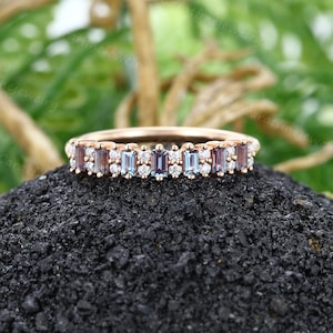May include: A rose gold band ring with a row of baguette-cut gemstones. The ring features a mix of clear, blue, and brown gemstones.