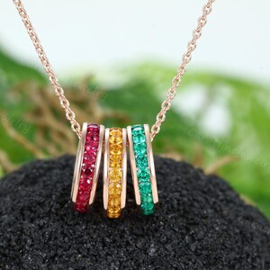 May include: A rose gold necklace with three rings, each set with a row of colorful gemstones. The rings are red, yellow, and green.