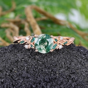 Könnte beinhalten: Ein Ring aus Roségold mit einem runden, facettierten Moosachat-Mittelstein mit grünen Einschlüssen. Die Ringschiene ist mit blattförmigen Akzenten verziert, die mit kleinen grünen Steinen und klaren runden Akzentsteinen besetzt sind.