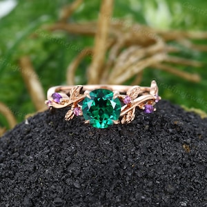 Op de afbeelding: Roségouden ring met een grote, ronde, smaragdgroene edelsteen. De band heeft blad- en takdetails met kleine roze en paarse edelstenen. De ring staat tegen een donkere, getextureerde achtergrond. Het ontwerp is geïnspireerd op de natuur.
