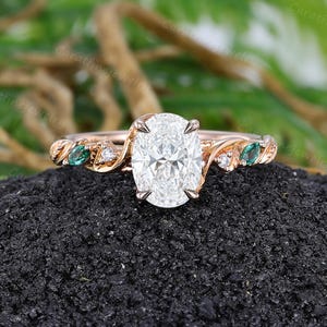 May include: A rose gold ring featuring a large oval-cut centre stone held by four prongs. The band has a twisted vine design accented with small round clear stones and marquise-cut green stones on both sides of the central setting.