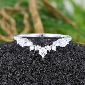 May include: A silver-toned curved band ring featuring a series of nine round-cut clear stones arranged in a V-shape. The stones vary in size, with the largest stone centred at the lowest point of the curve, flanked by smaller stones on each side.