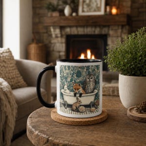 May include: A white ceramic mug with a black handle and rim, featuring a detailed illustration of a fox, an owl, and two rabbits in a clawfoot bathtub against a blue floral wallpaper background, resting on a round cork coaster.