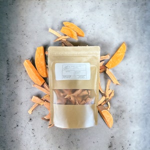 May include: A brown paper bag with a clear window showing sweet potato chews inside. The bag has a label that says "Sweet Potato Chews". There are several pieces of dried sweet potato scattered around the bag.