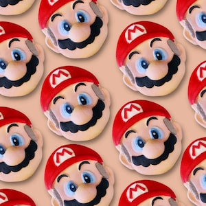 Könnte beinhalten: Ein Muster aus Mario-förmigen Badebomben. Jede Badebombe hat einen roten Hut mit einem weißen 'M', blaue Augen, einen schwarzen Schnurrbart und ein hellbraunes Gesicht. Der Hintergrund ist hellpfirsichfarben.