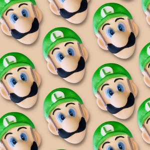 Puede incluir: Patrón repetido de bombas de baño con forma de Luigi. Cada bomba de baño presenta un gorro verde con una "L" blanca, ojos azules, un bigote negro y una cara bronceada. El fondo es de color beige claro.