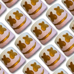 Könnte beinhalten: Ein sich wiederholendes Muster aus quadratischen Marshmallows mit einem lächelnden Gesicht. Jedes Marshmallow hat eine weiße Oberseite, einen rosa Körper und goldfarbene Gesichtsmerkmale. Das Bild zeigt ein sich wiederholendes Muster.