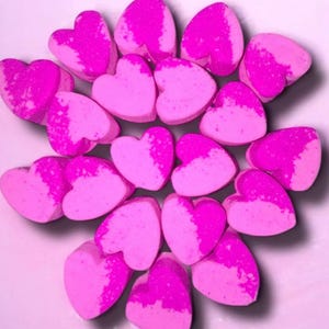 Op de afbeelding: Een verzameling hartvormige badbruisballen in roze en magenta tinten. Elke bruisbal heeft een tweekleurig ontwerp. De opstelling is cirkelvormig, met de harten in een radiaal patroon.