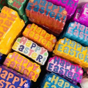 May include: A selection of brightly coloured bath bombs, each rectangular in shape. The bath bombs are in shades of pink, blue, yellow, orange, and green. The words "Happy Easter" are written on each in a contrasting colour.