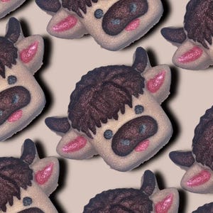 Puede incluir: Patrón repetido de bombas de baño con forma de vaca. Cada bomba de baño presenta una cara de color beige claro con flequillo, orejas y nariz de color marrón oscuro. Los ojos son azules y las mejillas rosas. El fondo es de color beige claro.