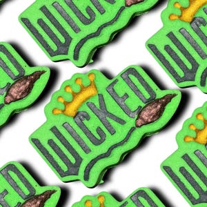 Puede incluir: Bombas de baño verdes en forma de corona con la palabra "WICKED" en letras gris oscuro. Cada bomba tiene una corona dorada y un acento marrón en forma de lágrima. El fondo es blanco.