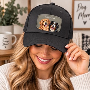 May include: A black trucker hat featuring a rectangular patch on the front. The patch displays a graphic illustration of a golden retriever and a Bernese mountain dog with the text Bentley & Cooper printed in white above the dogs.
