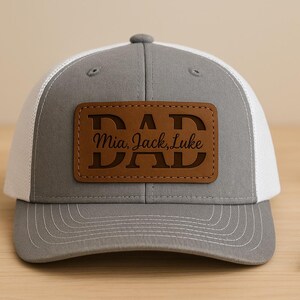 Op de afbeelding: Een grijze en witte truckerpet met een bruin leren patch. De patch leest "DAD" met de namen "Mia, Jack, Luke" eronder. De hoed staat op een houten oppervlak met een gouden ring en een pen.