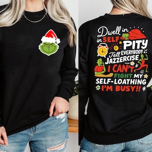 May include: Black long-sleeve sweatshirt featuring a Grinch graphic wearing a Santa hat on the front. The back of the sweatshirt has the text "Dwell in Self Pity" with other phrases and illustrations in a colorful design.