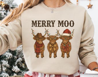 May include: Sand-colored sweatshirt with the text "MERRY MOO" above a graphic of three Highland cows decorated for Christmas. The cows wear a cowboy hat, a bow tie, and a Santa hat, with festive lights and scarves.