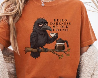 May include: Burnt orange t-shirt featuring a cartoon raven perched on a branch, holding a coffee cup and a coffee pot. The text "HELLO DARKNESS MY OLD FRIEND" is printed on the shirt. The shirt has short sleeves and a crew neck.
