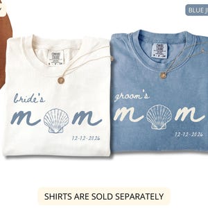 May include: Two t-shirts, one ivory and one blue jean, with the words "bride's mom" and "groom's mom" printed on them. Each shirt has a seashell graphic and the date "12-12-2026". Both shirts have a gold necklace.